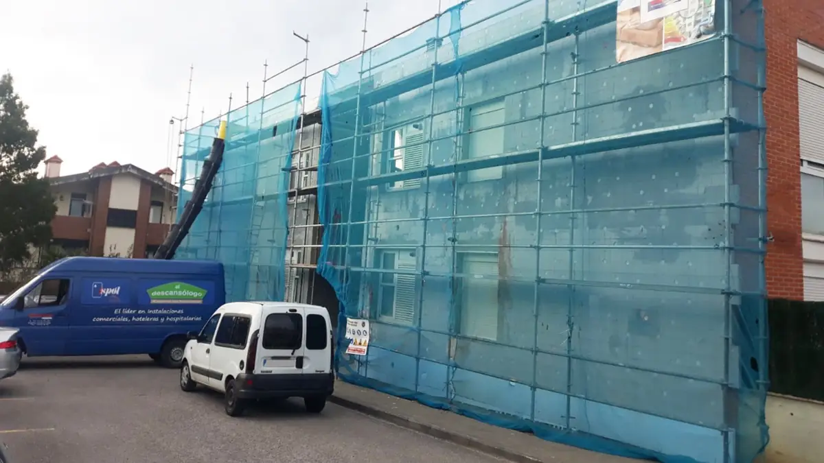 Fachada de un edificio con andamio y red de obra, con dos furgonetas de AM Decora delante